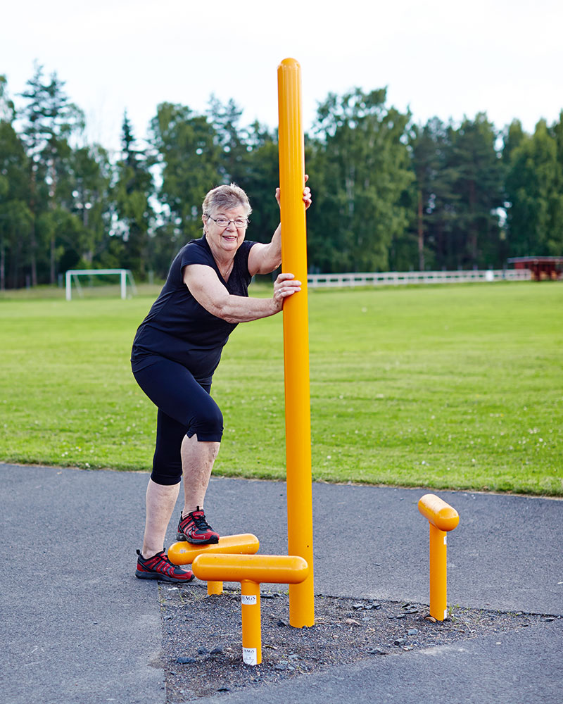 An elderly woman is completing the step up challenge at an outdoor gym station.